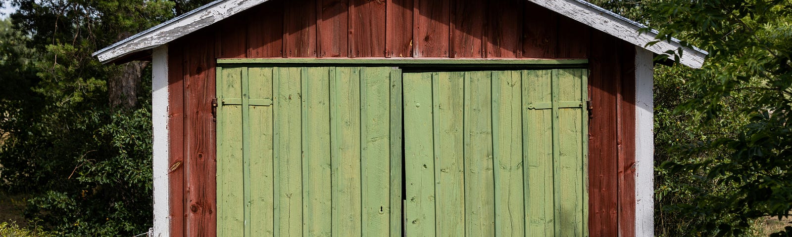 Brown wooden shed with light green double doors, surrounded by grass, with tall deciduous trees in the background.