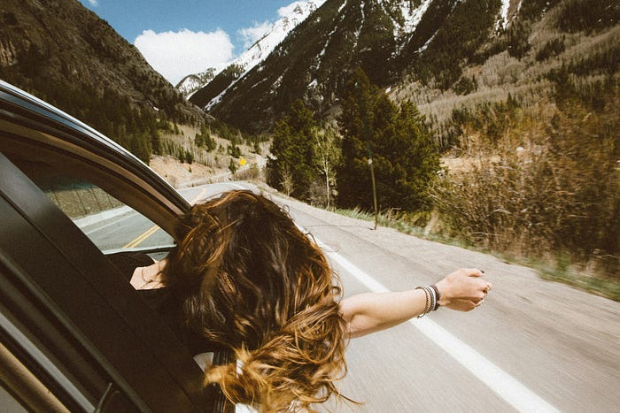 A woman with her head out of the car window, feeling the breeze, her hair blowing in the wind, while driving down a scenic mountain road lined with tall pine trees and snowy peaks in the background.