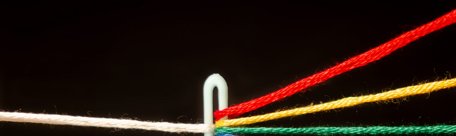 Close-up of eye of a needle, with red, yellow, green, blue and purple threads going in the eye, and a single white thread coming out of it.