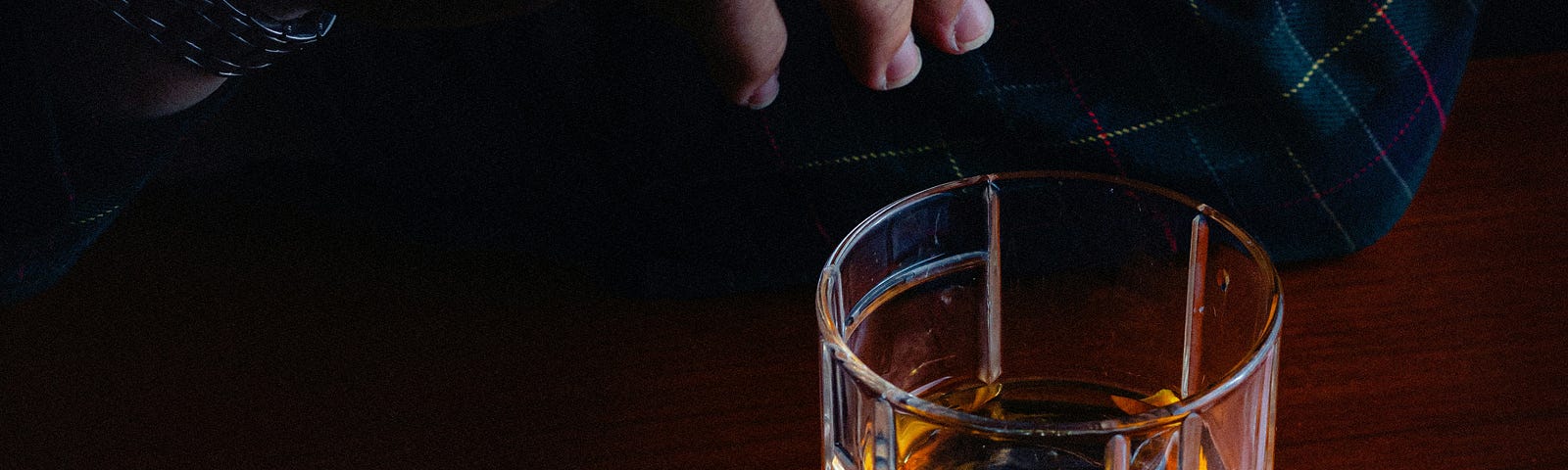 A man’s hand holding a cigarette over a glass of amber liquid at a bar.