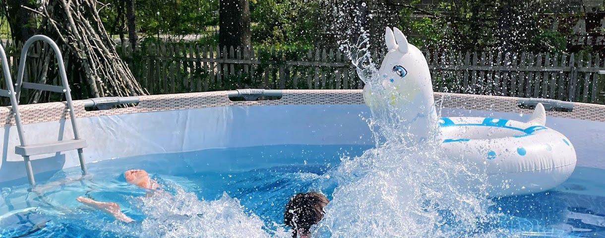 A person has just jumped into a swimming pool, but all you can see is a huge splash and the top of a head, with a pool float, ladder, and trees in the background.