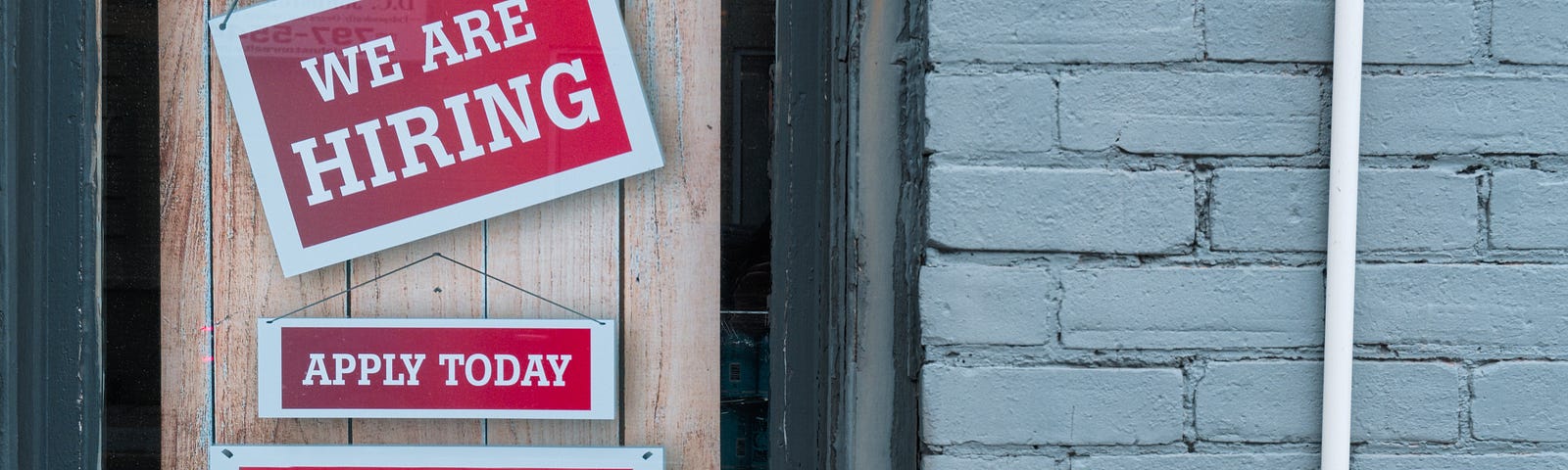 Red signs hanging on a door, saying “We’re Hiring” and “Apply Today.”