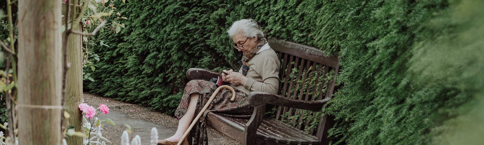 A senior woman is sitting alone on a bench and using her phone. It is a beautiful setting.