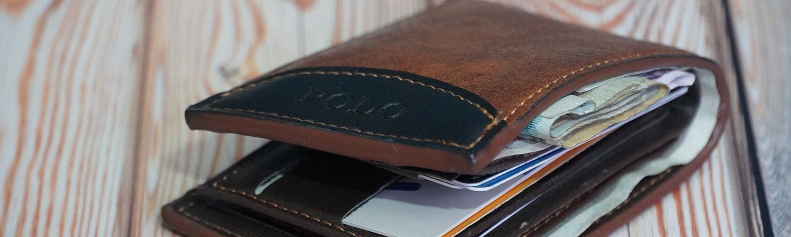 An image of a brown leather wallet with black trim on a rustic table. It has quite a few bills, cards and receipts in it — but it’s nowhere near as stuffed as mine is!