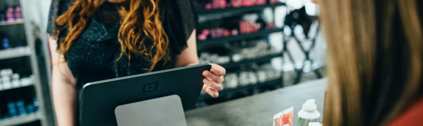 Two women, who’s faces you can not see, stand across from each other at a shop counter. One woman is using her credit card for a purchase.