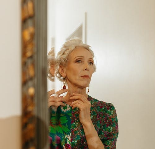 A well-dressed older woman contemplates her reflection in a mirror.