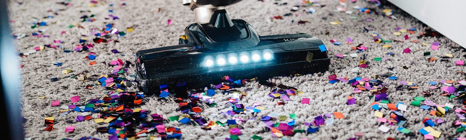 Image of a vacuum cleaner vacuuming many pieces of colorful confetti on a carpet.