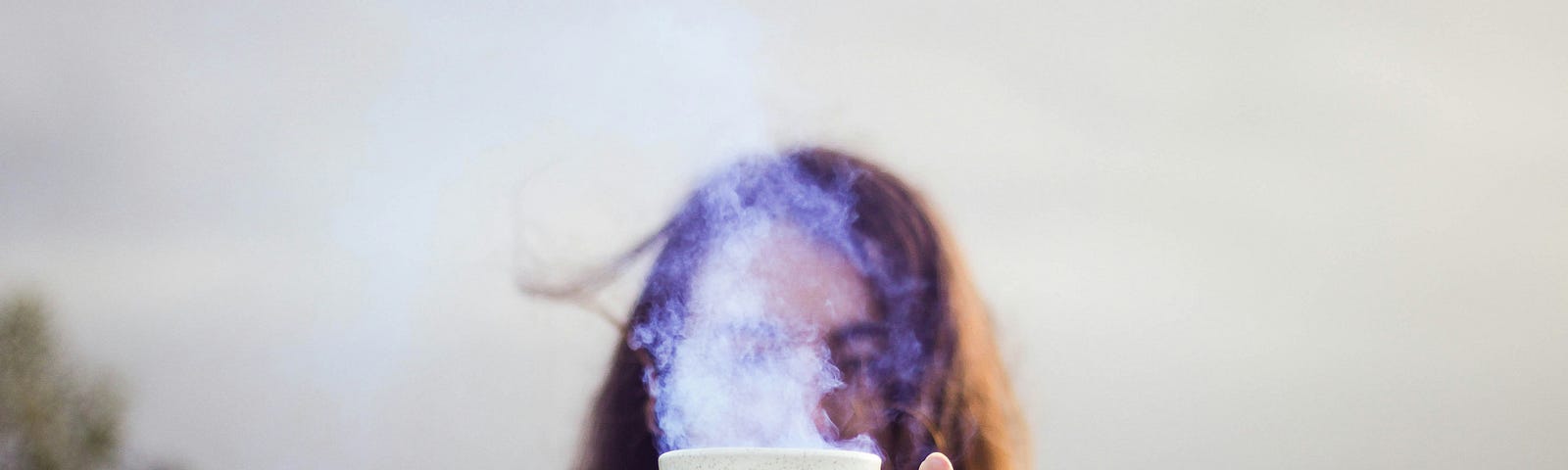 A person with long brown hair holds a cup of tea up to the camera, their face mysteriously obscured by the cup itself and the steam rising from it. The background is an out-of-focus rural scene.