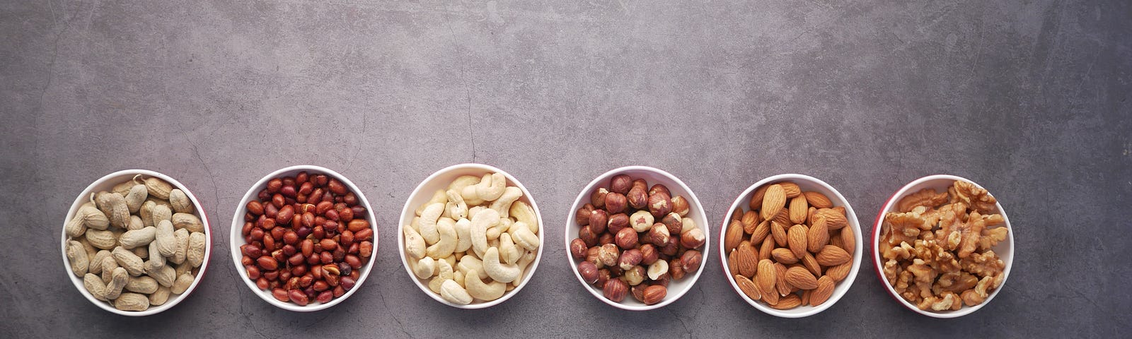 Six bowls of various nuts, lined up horizontally across the photograph. We have a bird’s eye view. The white bowls sit against  a gray background.