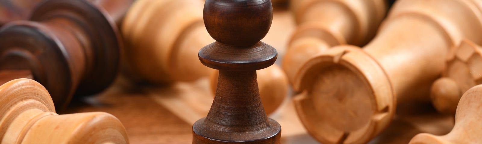 A lone pawn standing upright on a chess board, while all the other more valuable pieces are toppled over surrounding it.