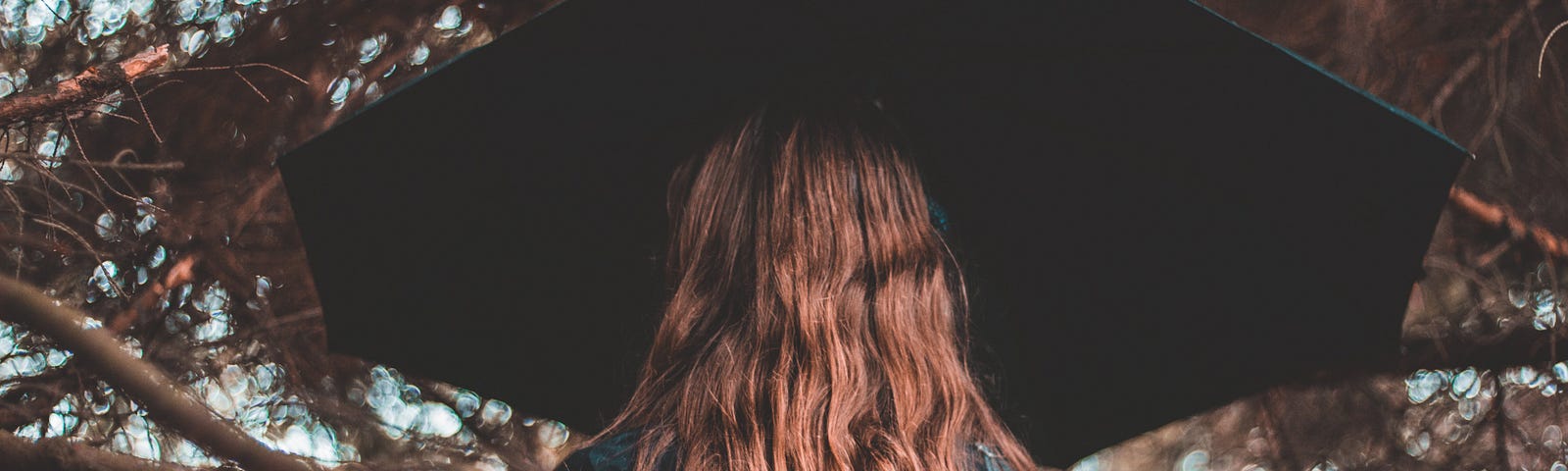 Long haired woman carrying a black umbrella standing under a tree, facing away from camera, some branches between the woman and the camera.