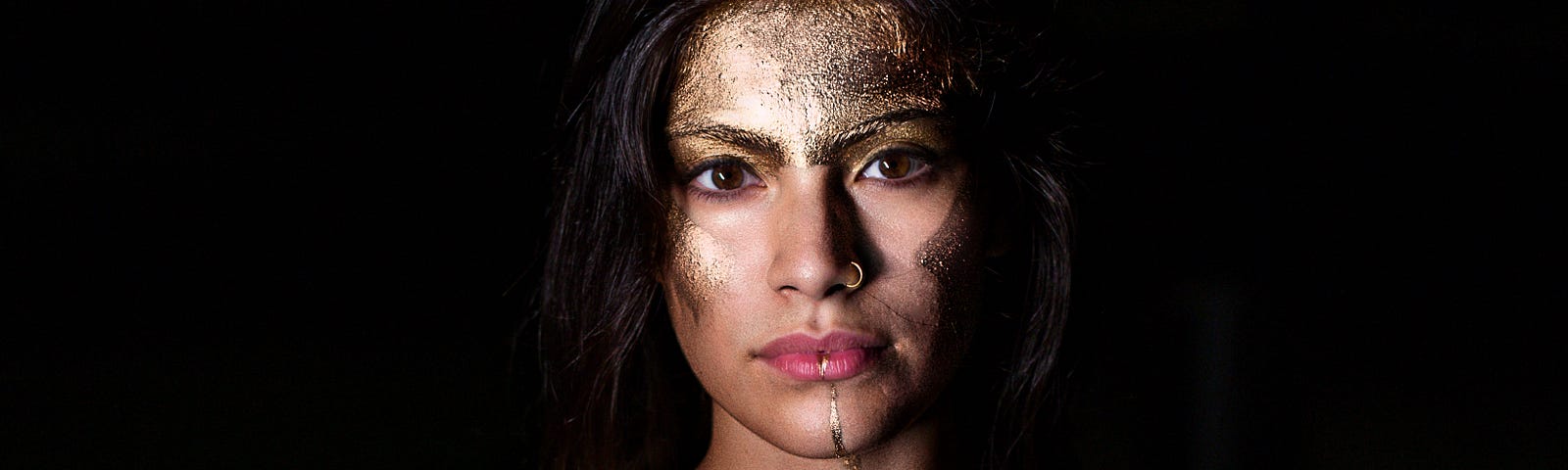 An image of a woman looking straight into the camera, and on her body she has golden glitter, accentuating her shoulders as if she’s wearing a top.