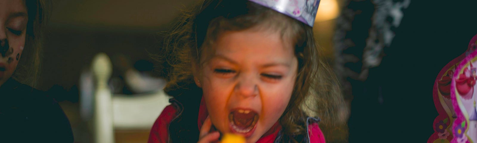 Picture of a young girl seated at a table in a party hat with her mouth wide open, either screaming or gathering breath to blow out 6 red candles on her birthday cake.