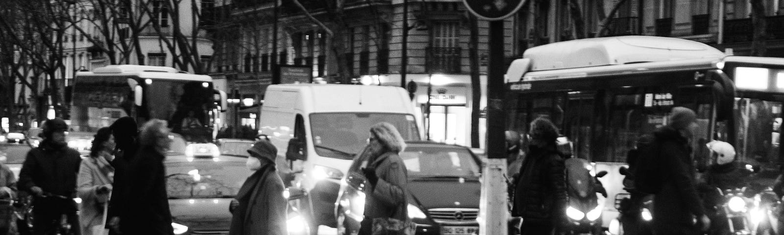 A black and white image of a busy street with people crossing it at a Zebra Zrossing and many vehicles waiting on the other side.