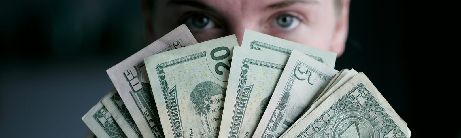 A person of an unknown gender holding 20 dolar bills in a fan in front of their face, obscuring the lower half. Only the eyes are visible over the edge.