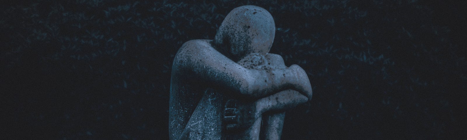 A blue stone statue sitting, legs drawn in, head on their knees as they weep in grief.