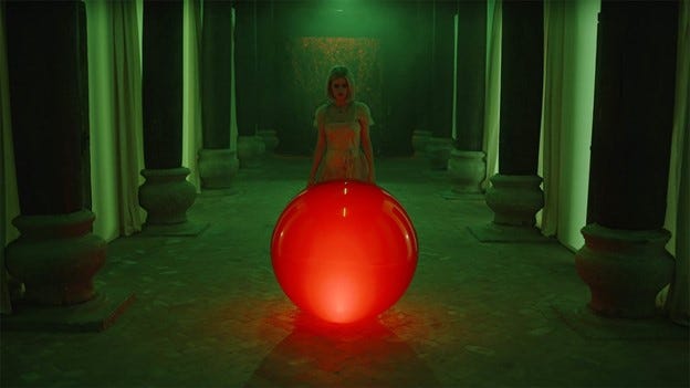 In an expansive, empty hallway lit in emerald green, lined with tall curtains and stone pillars, the young blond heroine approaches a glowing red orb reaching nearly to her waist.