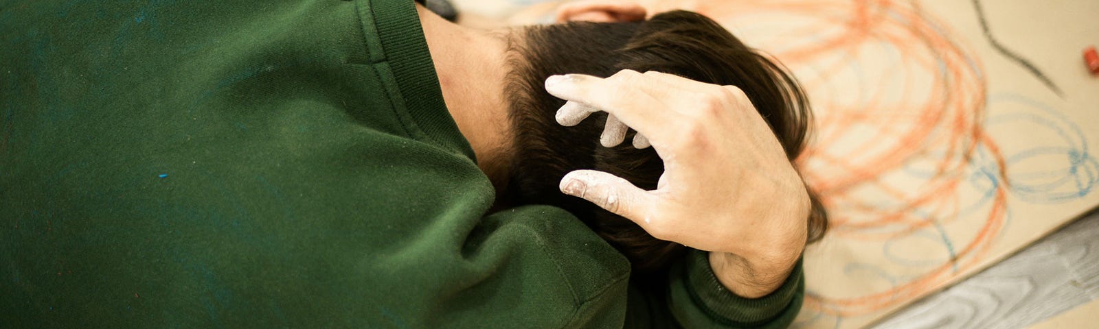 A person undergoing an art therapy exercise. Curled up on the floor wirth poaper and crayons, his hand over his head.