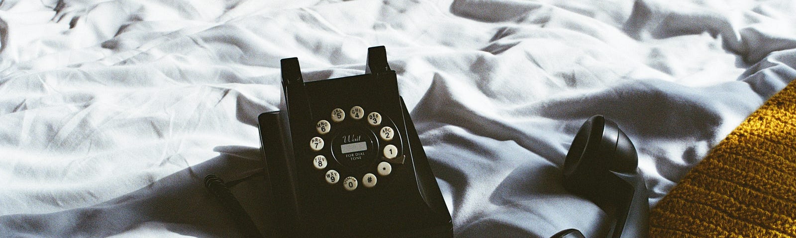A black dial phone sitting on a bed with the receiver unhooked