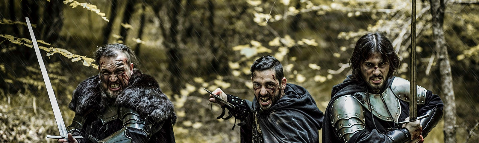 Three men in black mediaval armour with drawn swords and angry faces standing in a forest.