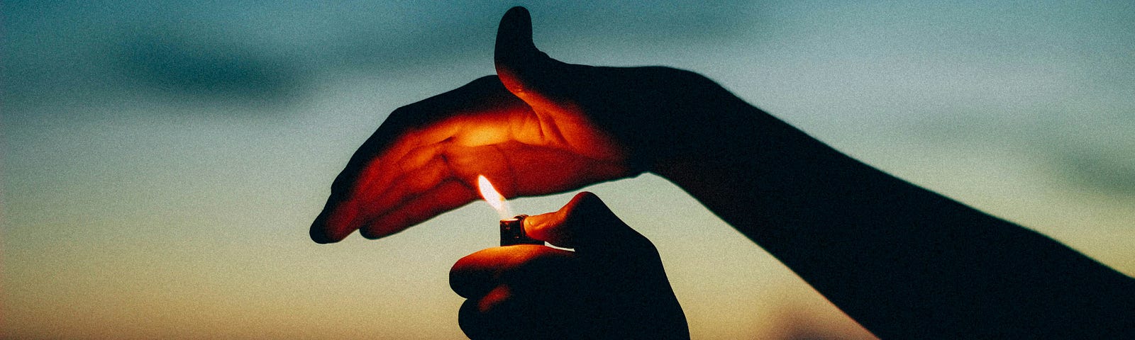 A hand holding a lighter with the flame burning against an open palm, outdoors, with the sun setting in the background.