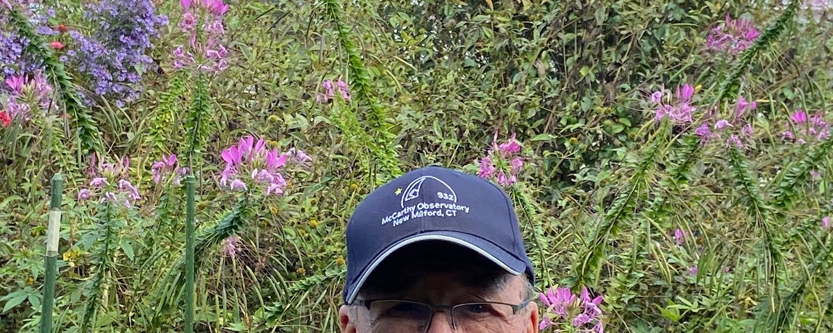 Bob Lambert, an older gentleman, wears a blue baseball cap and blue polo shirt, and stands in front of native flowers at the observatory.