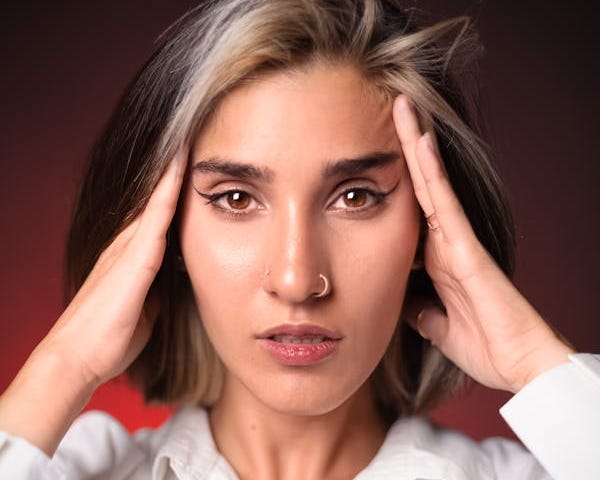 Profile of a woman with highlights and a nose ring in a smart white shirt staring into the camera and rubbing her temples in frustration in front of a deep and dark red background