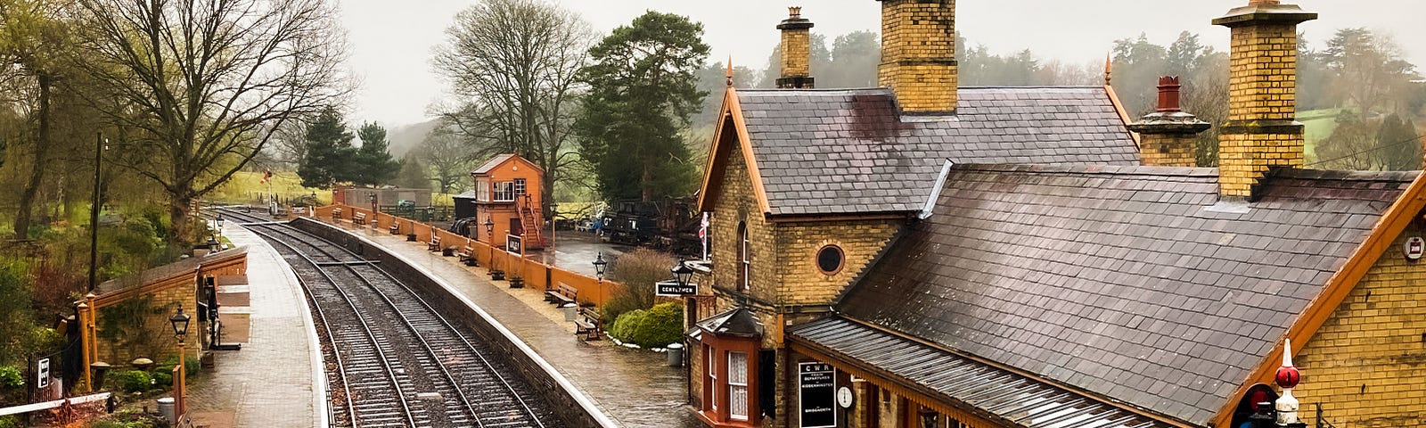 Image of a small suburban station and platform seen from the air, the train tracks on the left and the station building on the right.