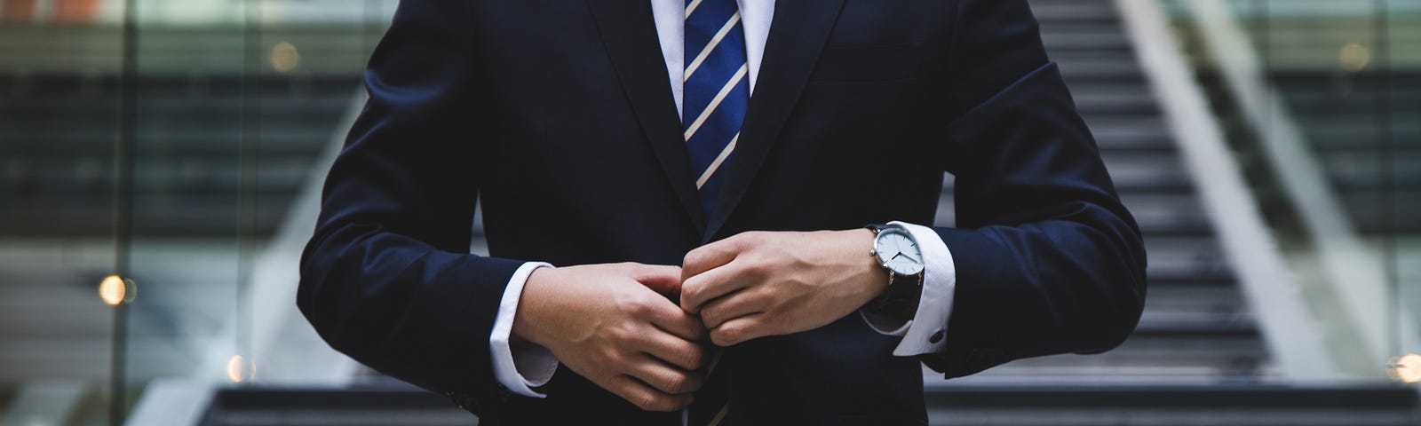 The torso of a man in a navy three button suit. He is buttoning the top button.