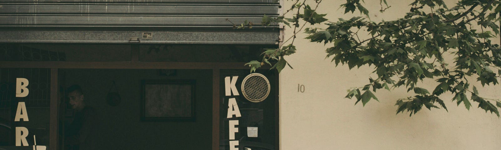 Two older men are sitting outside a small café, engaged in conversation at a table under a tree.