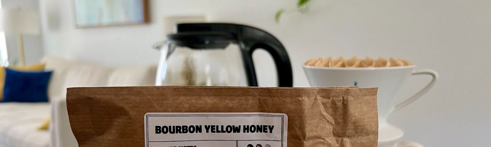 A hand holds up a bag of coffee labeled “Bourbon Yellow Honey.” In the background is a coffee mug, coffee pot, and furniture.