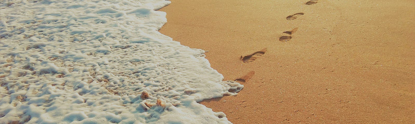 Surf foams washing over a set of footprints on a sandy beach.