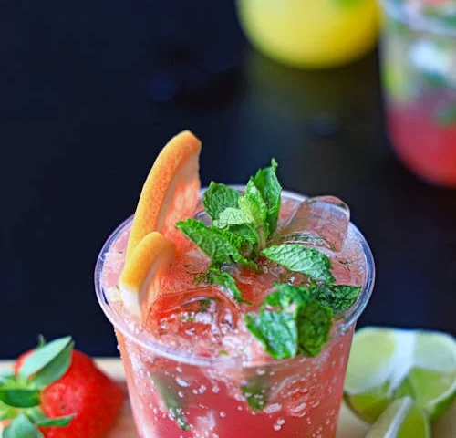 A photograph of dark pink punch garnished with mint leaves, orange and lemon slices. It is placed on a light brown wooden cutting board with a lime cut in half, a wedge of lime, a strawberry, mint leaves, and other garnishes.