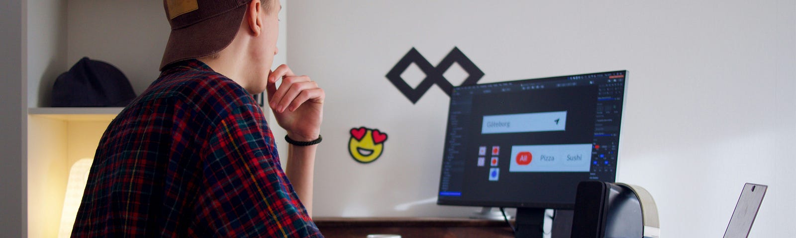 A young freelancer wearing a hat looking at his desktop screen with a laptop on the side