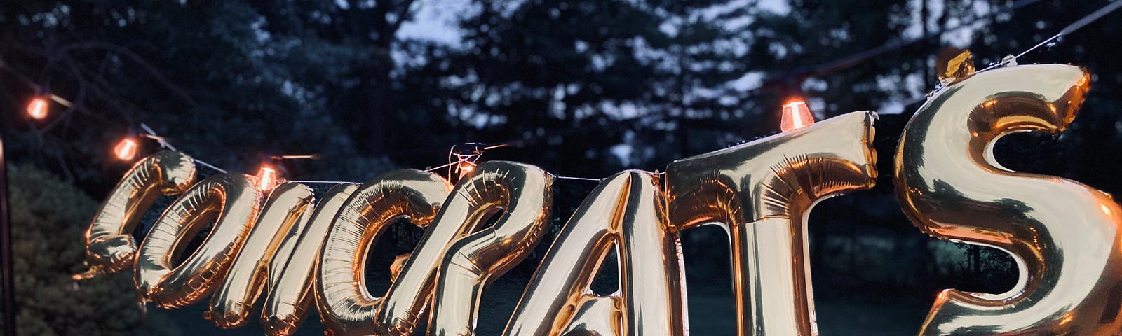 A congratulations balloon sign hanging between 2 trees.