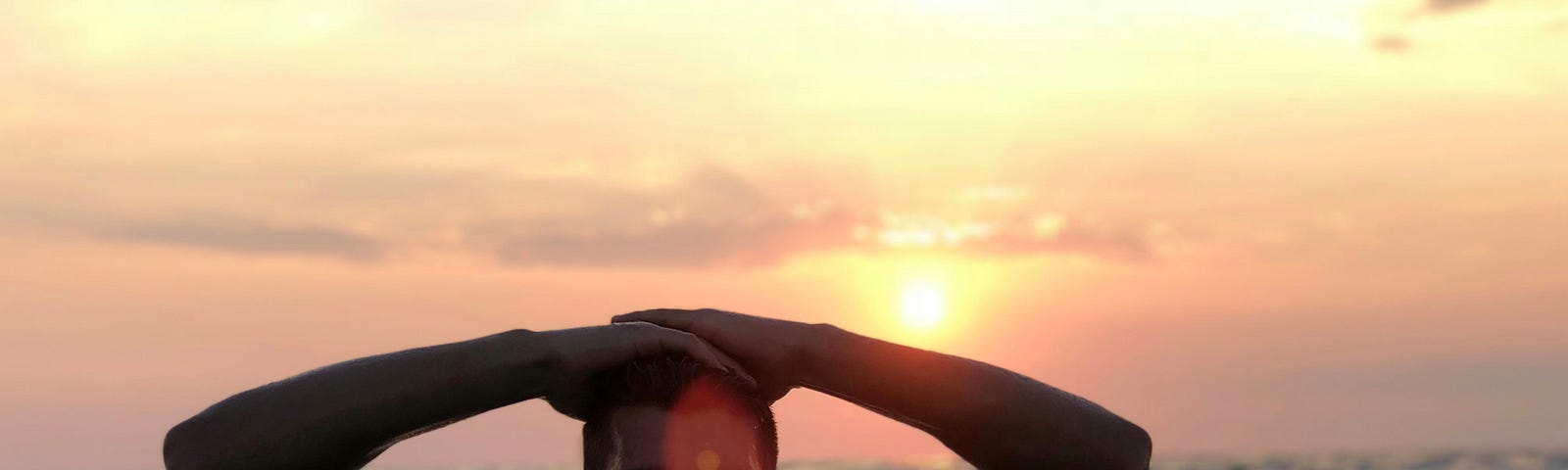 A muscular man stands in the oceanm his hands on his head, The sun sets in the background.