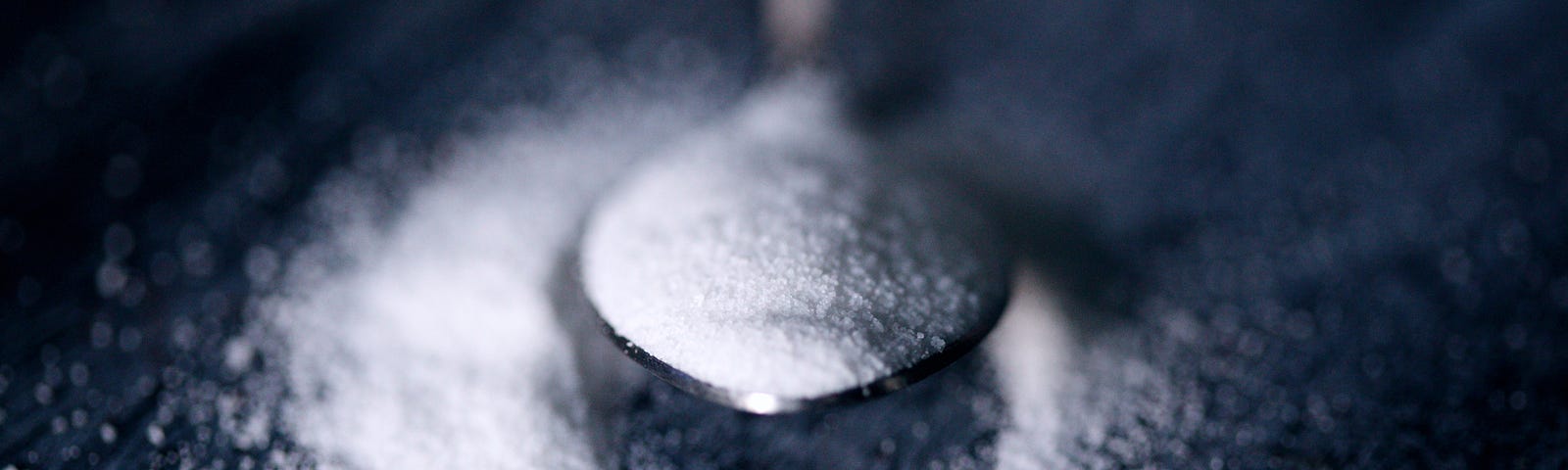 A silver narrow, heart-shaped spoon emerges from the image top, with sugar spilling over to create a large white circle on a deep blue background. A recent review published in the British Medical Journal reminds us that excessive sugar consumption is associated with the following health problems: Weight gain; type 2 diabetes; gout; and cancer (certain kinds).