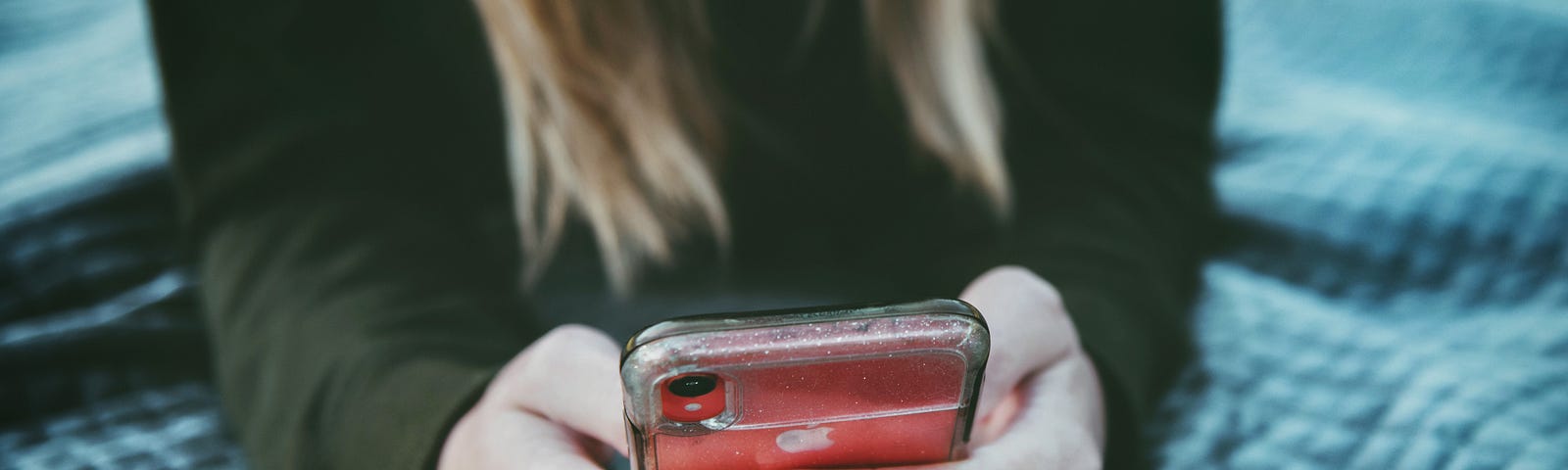 closeup of a person’s hands holding a red cell phone. You can also tell that this person has long black sleeves, is sitting on a blue quilt and has long blond hair. The face and neck are cut off.