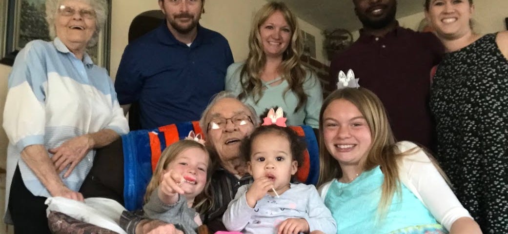 A older gentleman sits in a recliner with his three great granddaughters on his lap. Behind him stand his granchildren and their spouses.