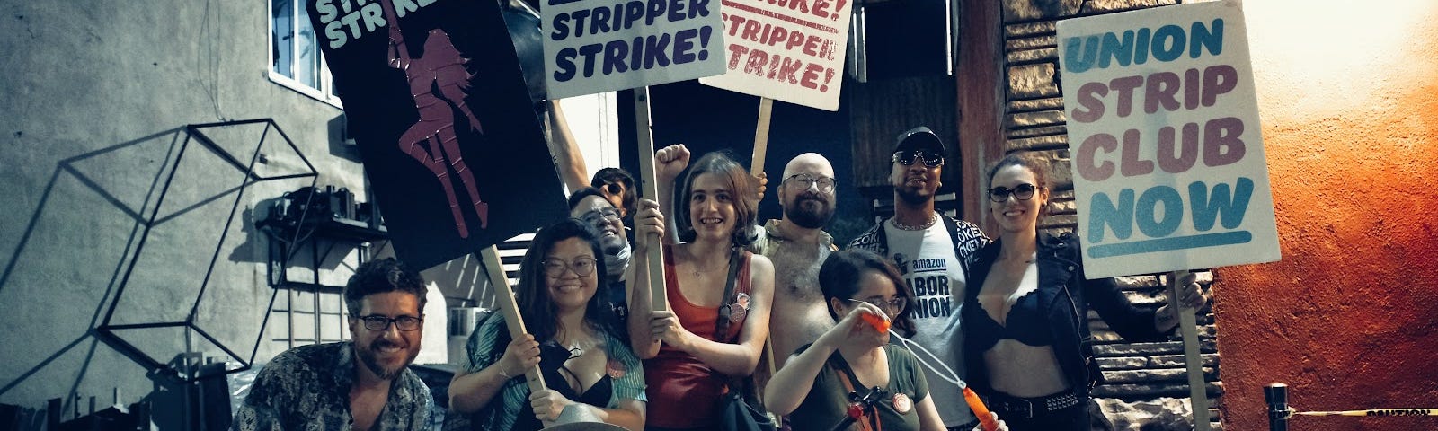 A group of video game workers and allies pose for a photo in front of the Star Garden Dive Bar. Workers hold colorful signs reading “STRIPPER STRIKE” and “UNION STRIP CLUB NOW.” Amazon Labor Union president Chris Smalls is also present