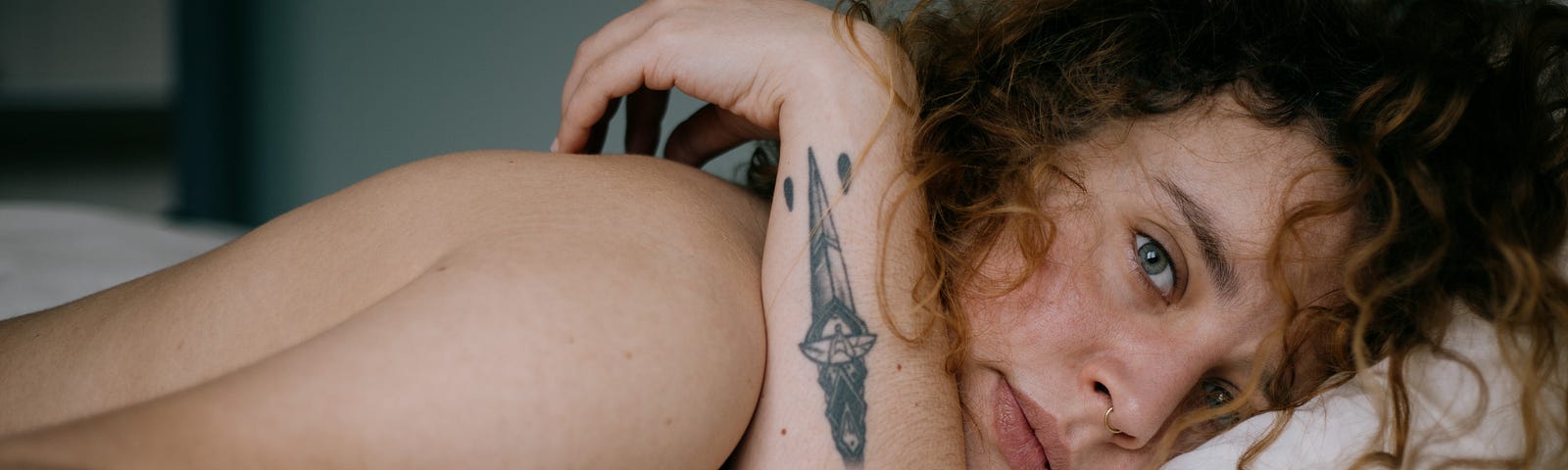 woman lies in bed, one arm up showing dagger tattoo