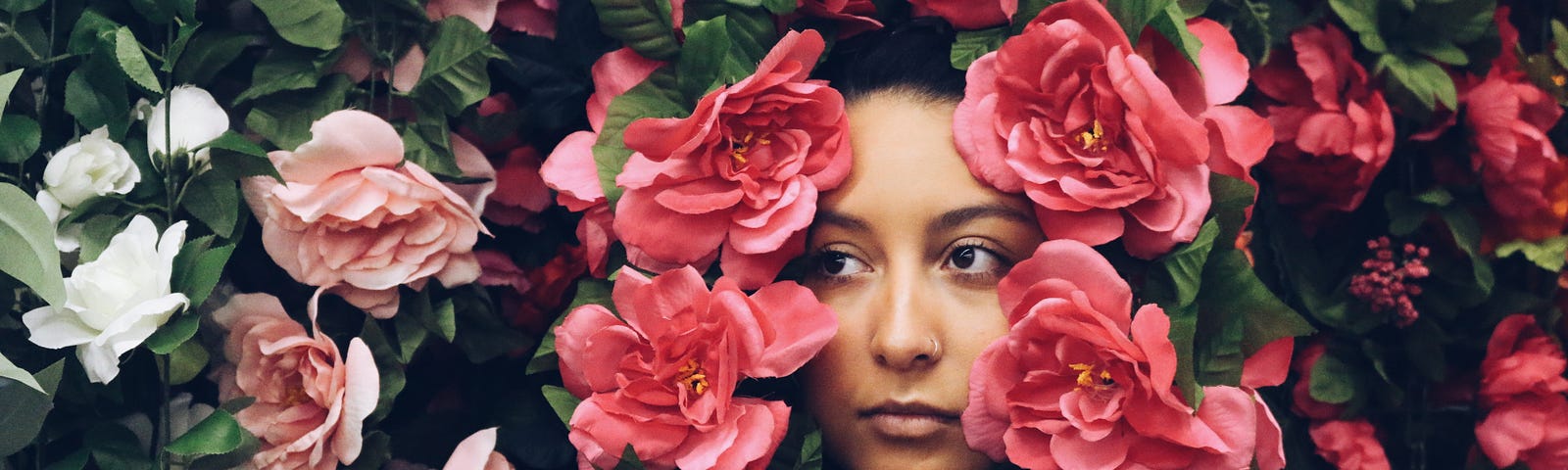 A woman stands amongst a wall of flowers, with the flowers cascading around her face. She looks thoughtful.