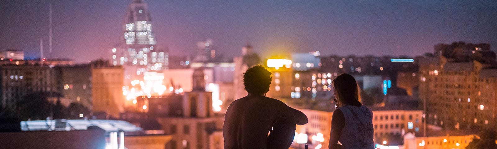 A man and a woman sit on a rooftop ledge, overlooking a city lit up in the evening