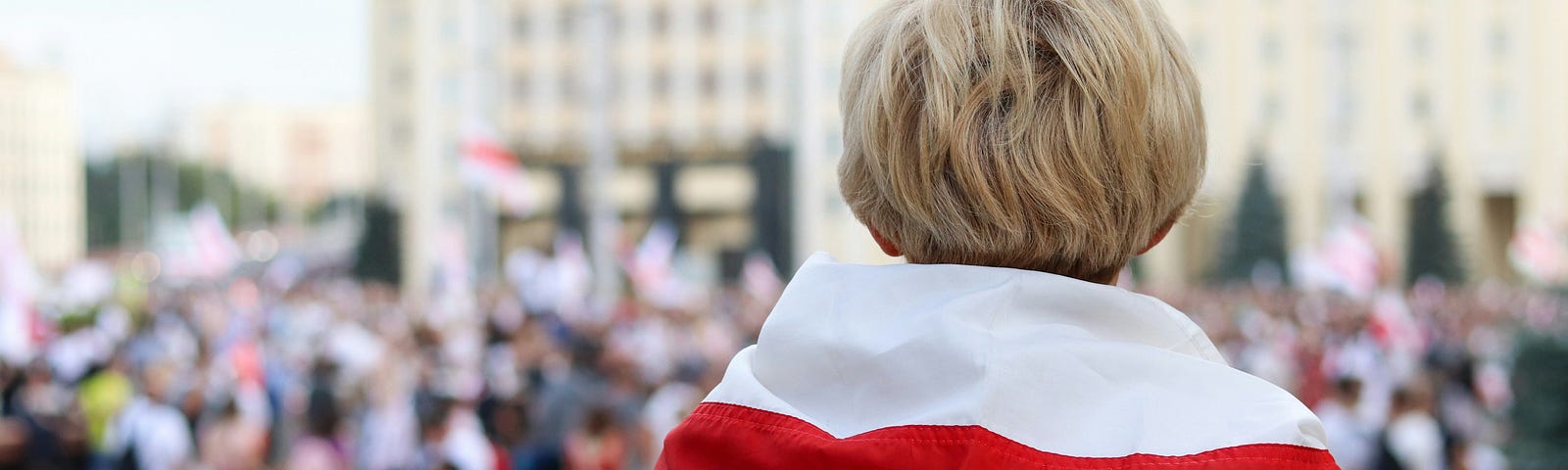 A person draped in a red and white flag observes a group of demonstrators in a city square, from a distance. The demonstrators are out of focus — only the observer, viewed from behind, is sharply focused.