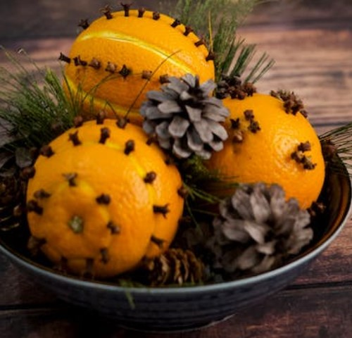 A photograph of a green ceramic bowl filled with orange pomanders, pine cones, and greenery sitting on a wooden surface.