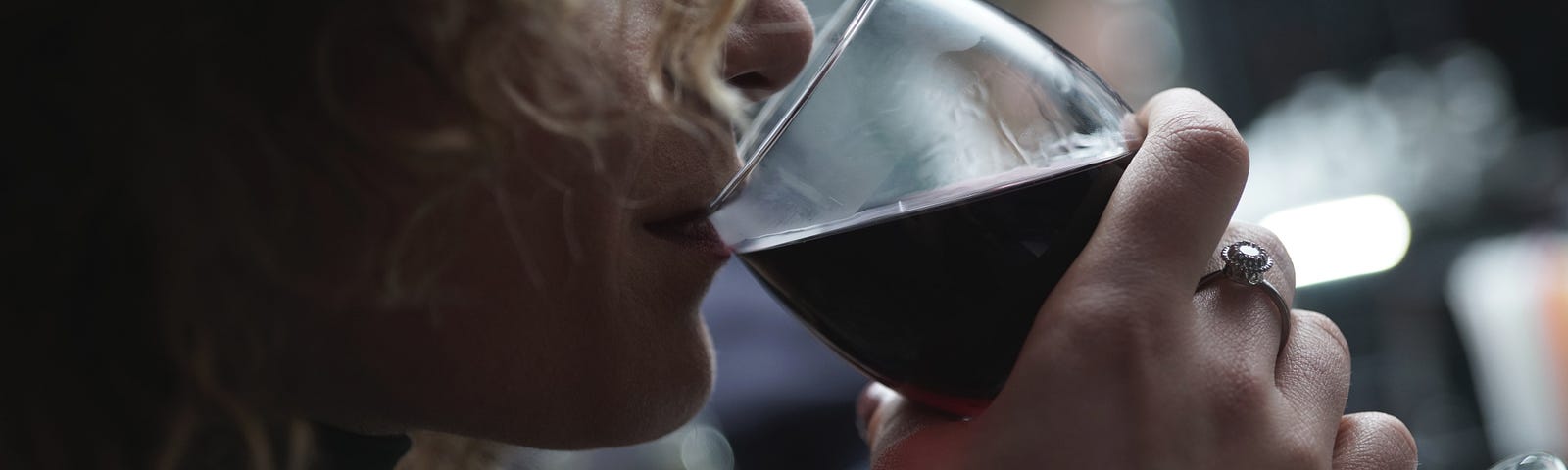 A close-up profile of a person with long curly hair hiding their face drinking red wine from a stemmed glass.