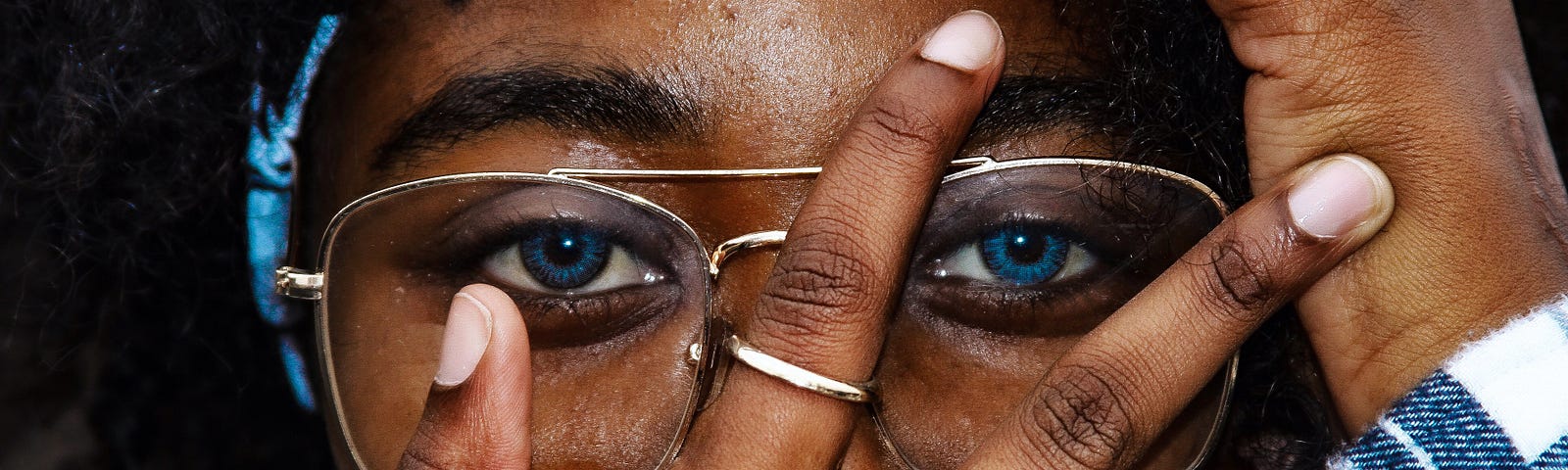 A close-up picture of a woman wearing glasses. Her right hand is spread over her face with the thumb pointed at her right eye. The right index and middle fingers are make a V shape over the left eye. Her left hand is supporting the left side of her face.