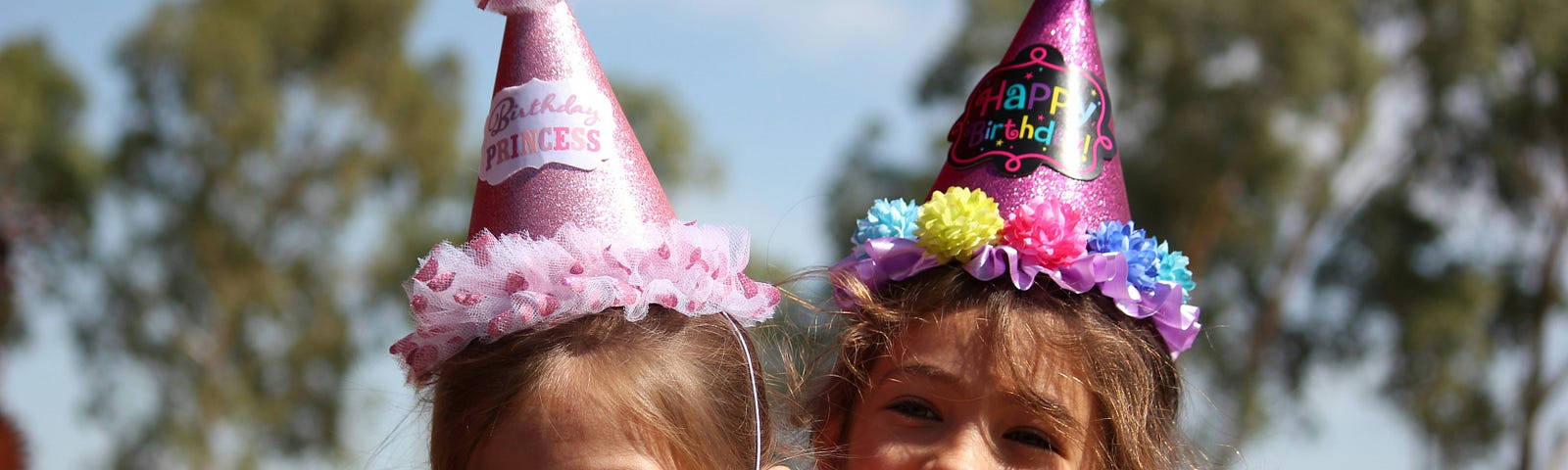 Two happy children at a birthday party. Both girls are wearing a party hat with a pom pom on the top. One hat says, “Birthday Princess” and the other says, “Happy Birthday.”