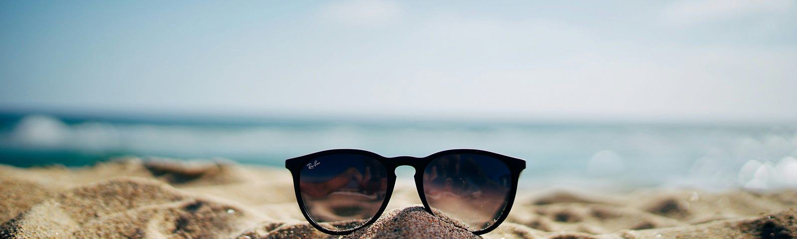 A pair of sunglasses sittin in the sand, sparkling sea, and blue sky behind.