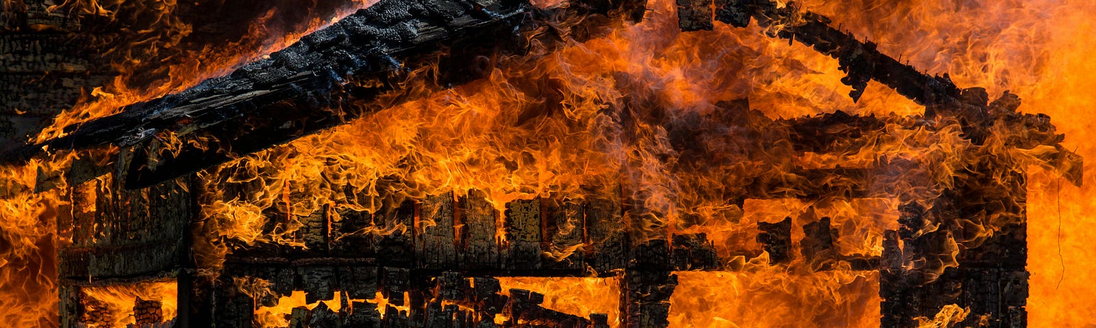 Image of a building on fire; it might have been a cabin. The only thing left and still ablaze is the framework, which appears blackened and ready to collapse. The flames are bright orange and seem out of control. Some dark grey smoke is also visible in the shot.